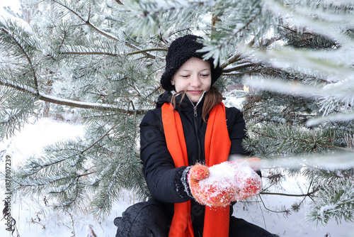 A thirteen-year-old girl wearing a black and bright orange scarf and mittens holds snow in her hands against a backdrop of trees on a winter day