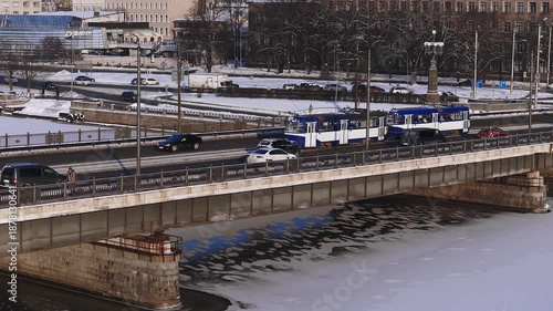 Aerial 4K winter scene in Riga, Latvia shows the Daugava River, a stone and steel road and tram bridge, blue and white trams, steady traffic, and historic church spires.