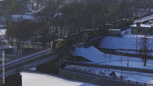 Aerial view of a yellow and dark commuter train crossing a steel railway bridge in Riga, Latvia, over a frozen canal, with cars below, leafless trees, and chimney smoke.