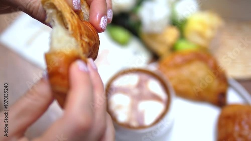 Female hands with a sparkling manicure breaking a flaky, buttery croissant in half, showing the soft inside, with a cup of hot chocolate and other pastries blurred in the background