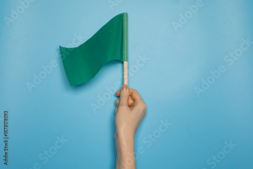 Hand holding a green flag against a blue background. Concept of permission, approval, safety, positive signal, start or go ahead symbol with copy space.