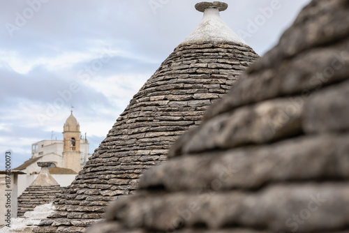 фотографія conical limestone roofs of traditional trulli houses in alberobello, apulia, italy, featuring stacked stones, a cloudy sky, and a historic church bell tower