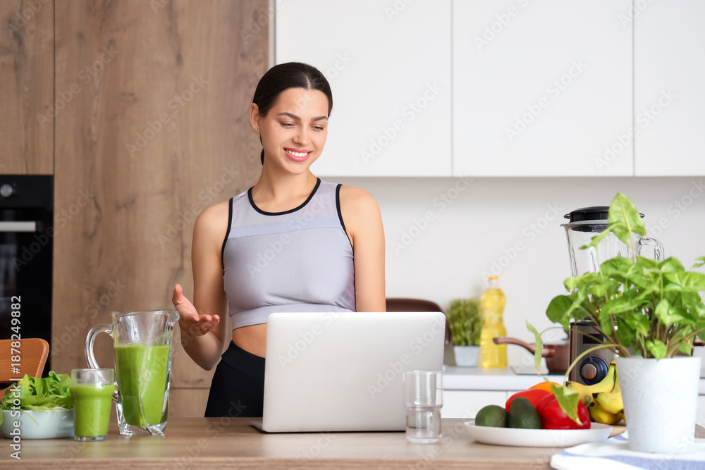 Fototapeta premium Sporty young woman with healthy smoothie and laptop in kitchen