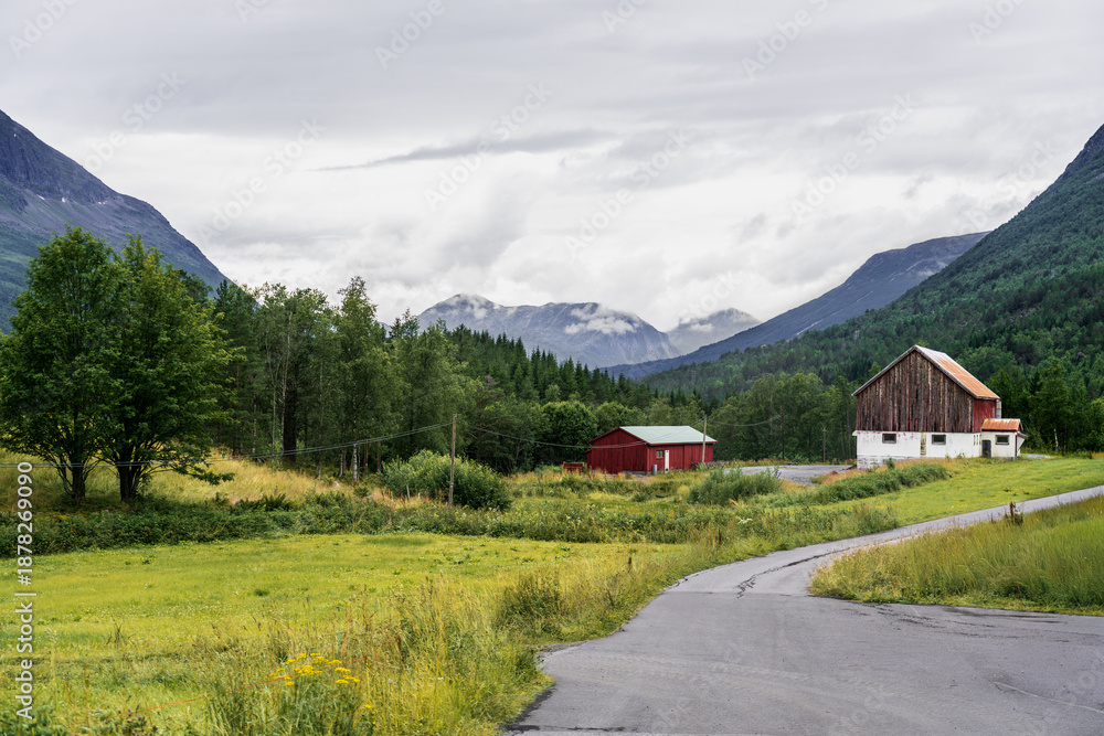 Obraz premium A barn stands beside a winding road in Innerdalen. The green valley is surrounded by mountains and clouds, showing the natural beauty of Norway's landscape
