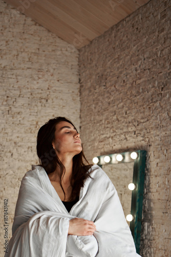 A woman with black hair wakes up in the morning in a white blanket and catches the rays of the sun. The portrait is creative, with brown eyes. High quality photo