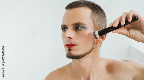 portrait of gay man with makeup and cosmetics on face. A handsome young transgender man with red lipstick on lips and painted eyes applies blush with a brush and does facial contouring