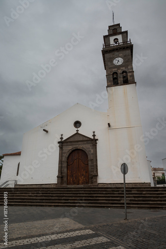 Nuestra Señora de los Remedios, a white church with a tall bell tower and stone entrance in Buenavista del Norte, Tenerife, Spain, stands under a cloudy sky