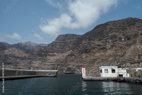 View of the entrance to the marina of Santiago del Teide, Tenerife, Spain, with the imposing Los Gigantes cliffs rising dramatically in the background under a bright blue sky