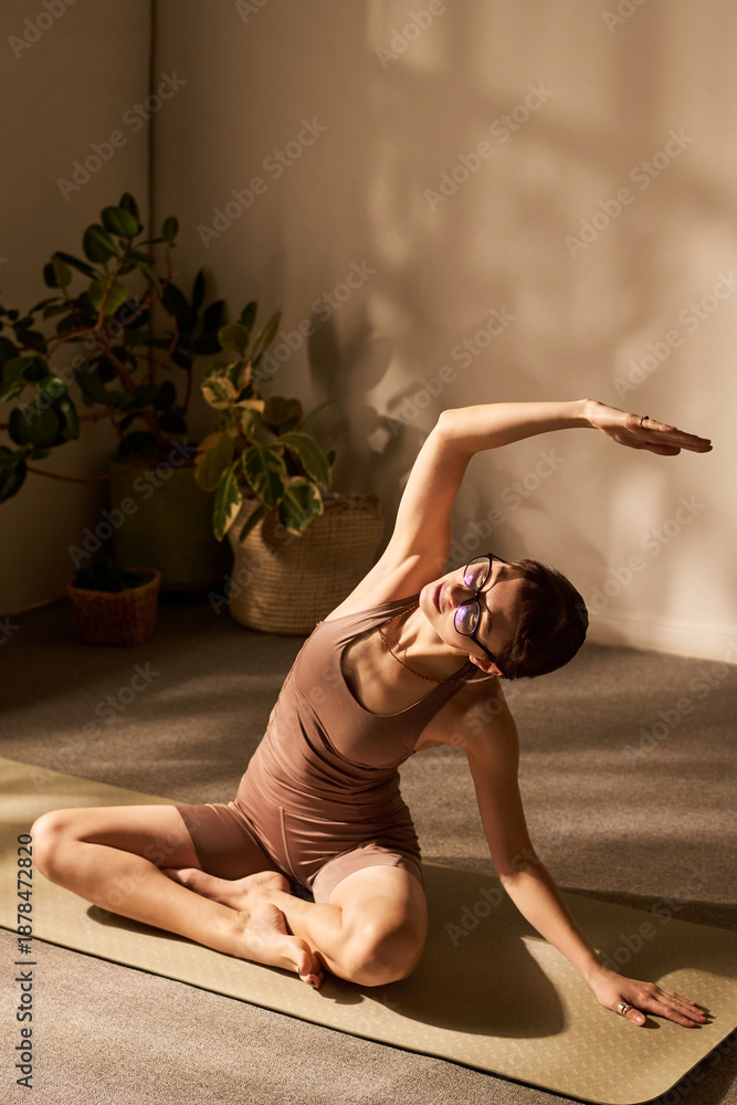 Fototapeta premium Caucasian young adult woman sitting on yoga mat performing seated side stretch with arm raised, wearing glasses, practicing yoga or pilates in bright indoor space