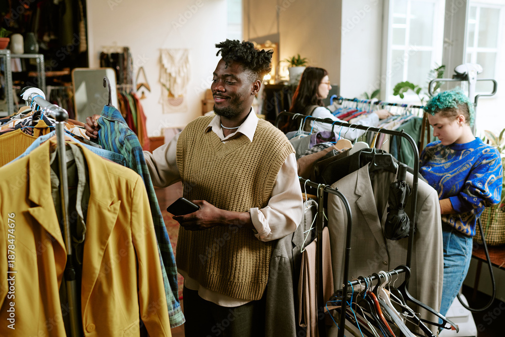 Fototapeta premium Young Black man browsing clothing racks in second hand store, holding smartphone, standing among diverse group of shoppers including young Caucasian woman with green hair searching for used clothes