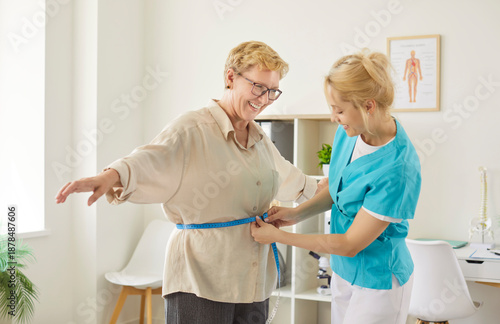 Obraz na plátně Nurse measuring waist of smiling senior woman with tape to check BMI on medical checkup in clinic