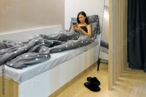 A woman is laying in bed and looking at her phone. She is covered in a grey blanket with white triangles
