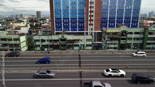 Slow motion footage of multi-lane expressway with vehicles passing weathered apartment complexes meters from road, contrasted by modern blue high-rise in San Andres, Manila.