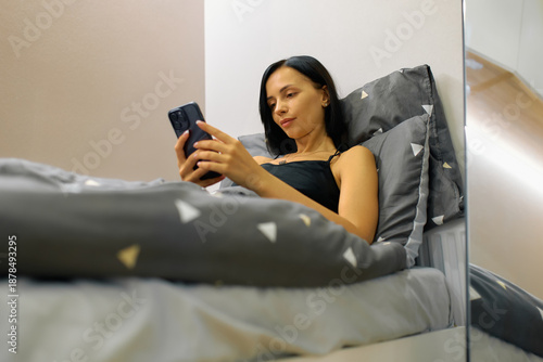 A woman with long dark hair is laying in bed and looking at her phone