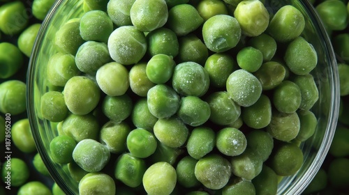A clear bowl holds frozen green peas. Surrounding the bowl is a larger group of green peas placed together. The scene takes place in a kitchen during daylight. © mila103
