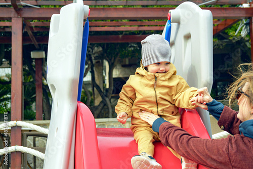 Bild auf Leinwand Joyful toddler sliding down colorful playground with mother's supportive hand nearby