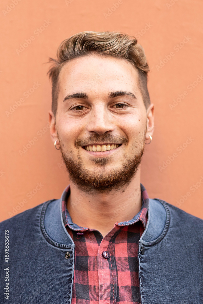 Obraz premium Smiling young man standing in front of a wall wearing a checkered shirt and denim jacket. He looks confident, friendly and approachable. Millennial generation and modern casual style concept.