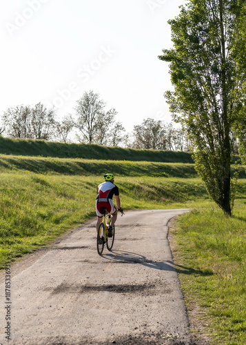 Cyclist riding alone on a country road surrounded by green fields and trees during a sunny day. He wears professional cycling gear and a yellow helmet. Outdoor activity and healthy lifestyle concept.