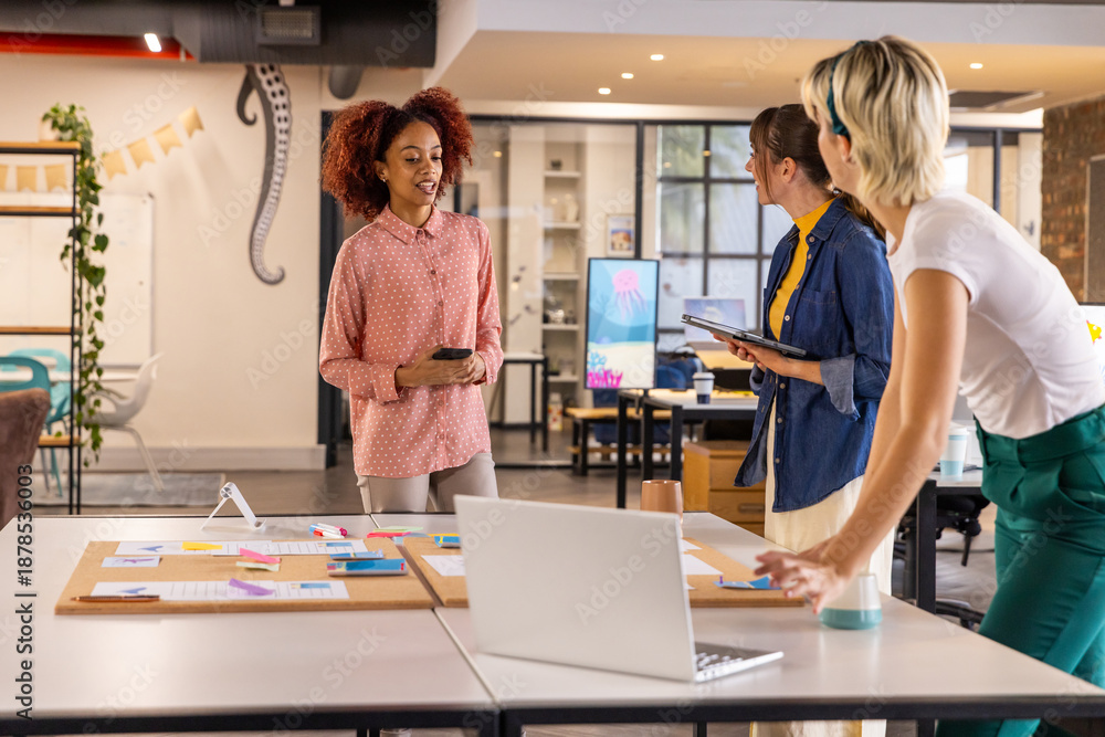 Obraz premium Diverse female colleagues collaborating around table in office holding smartphone, tablet