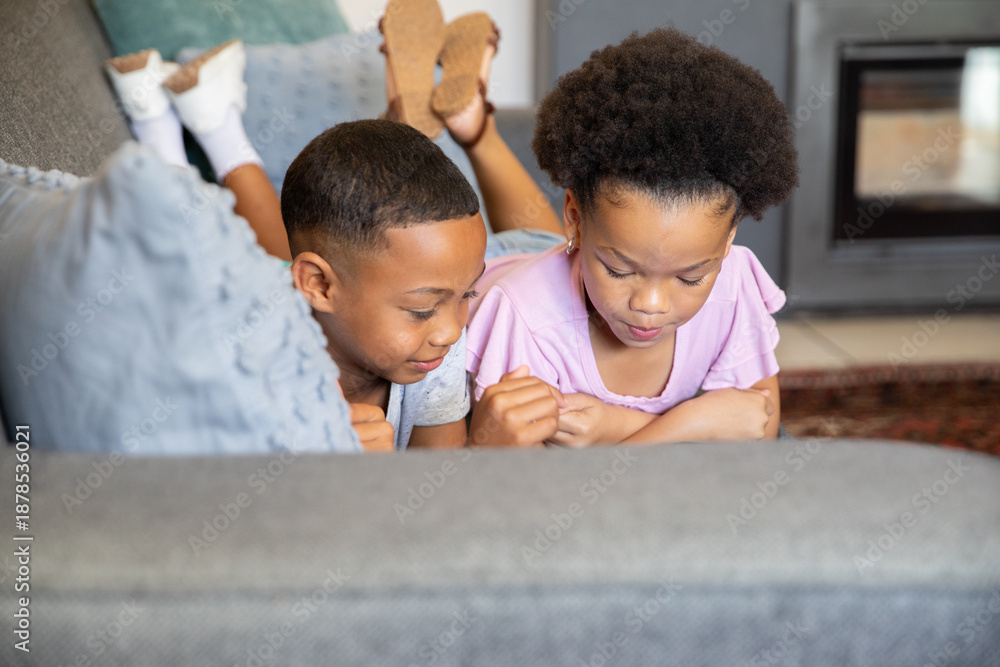 Fototapeta premium Diverse child siblings lying on grey sofa in living room looking down near light blue pillow