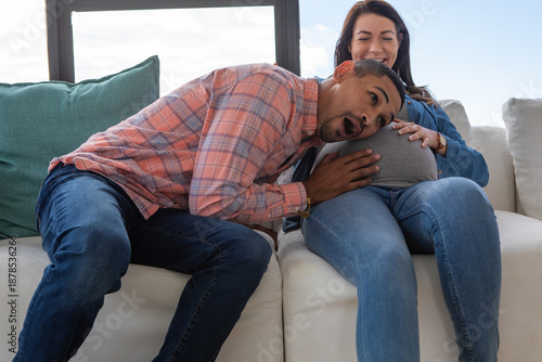 Couple leaning in, pressing ear against baby bump on white sofa with cushions by window