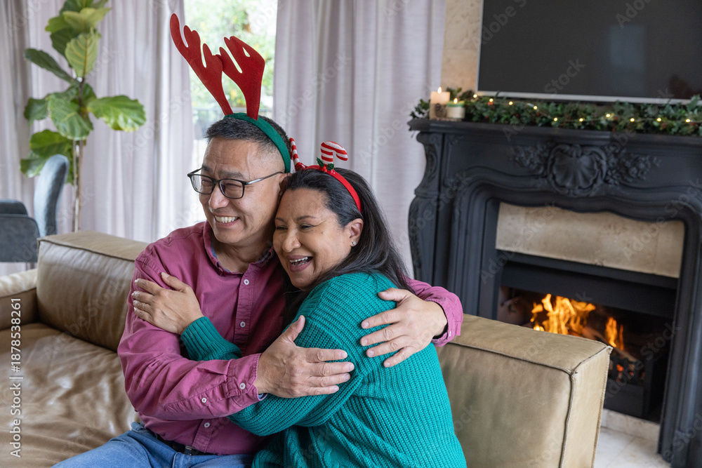 Fototapeta premium Senior couple hugging while sitting on couch in living room with lit fireplace, antler headbands