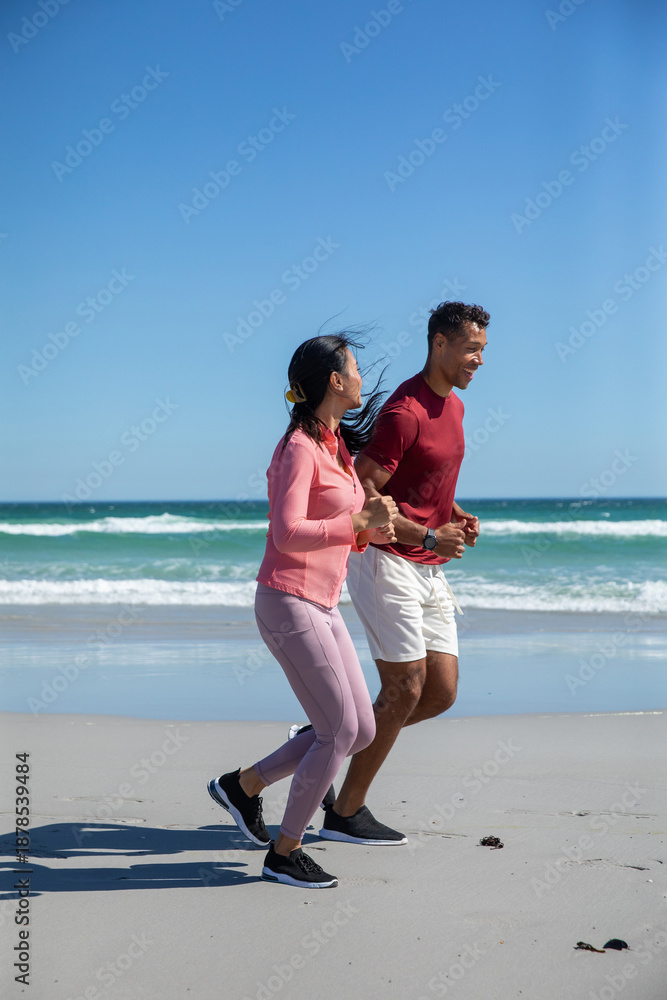 Fototapeta premium Diverse couple jogging along sandy beach shoreline in athletic clothing and running shoes