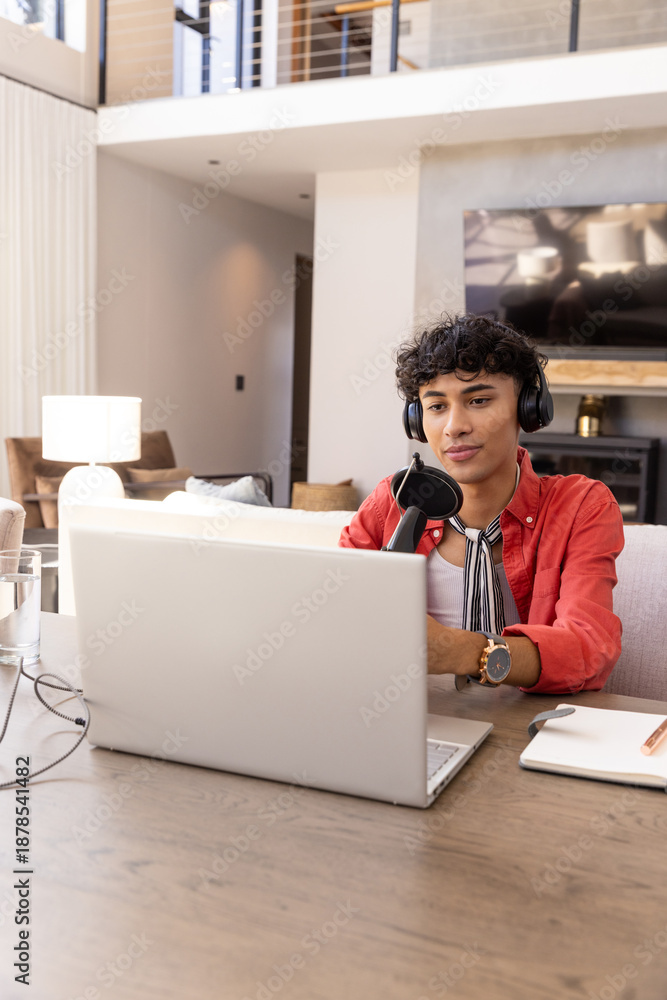 Naklejka premium Male broadcaster wearing headphones sitting at wooden desk in living room using laptop