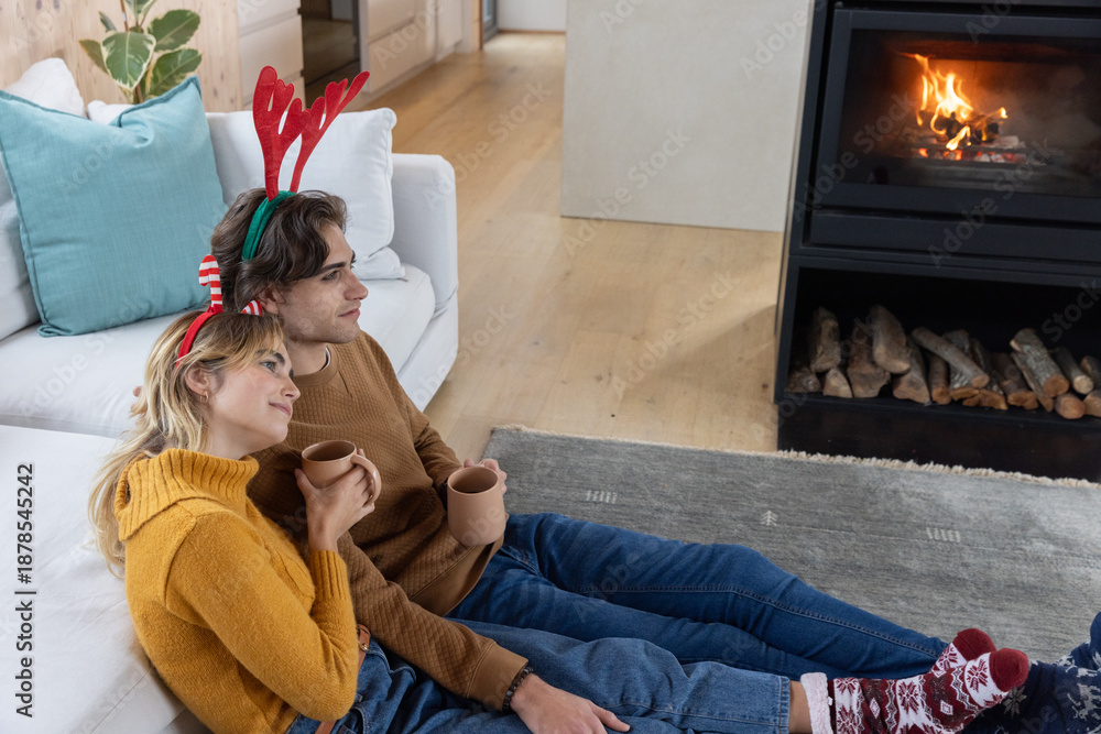 Fototapeta premium Couple wearing red antler headbands leaning against sofa, holding mugs at home before fireplace