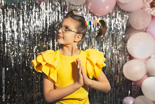 Happy funny girl in glasses and yellow holiday blouse ponytails hairstyle with hands folded in prayer on festive background