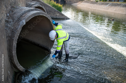 A male resource worker is collecting water samples flowing from a large drainage pipe.