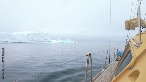 Side view of sailing boat passes massive iceberg in foggy Arctic waters