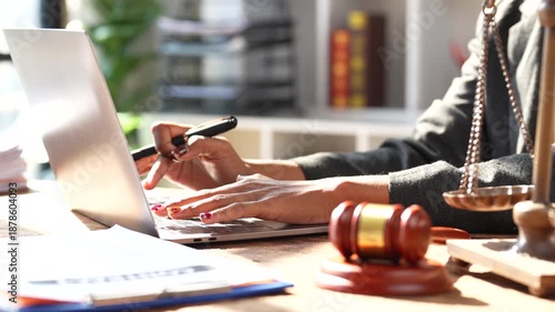 Professional african american female lawyer working on a laptop at her desk with a gavel and scales of justice, focusing on a case, legal advice, and courtroom preparation in a modern law firm