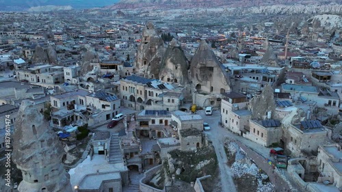 Aerial drone footage of Göreme town in Cappadocia, Turkey, surrounded by fairy chimneys and volcanic rock formations. The shot reveals traditional stone houses, cave dwellings carved into soft rocks.