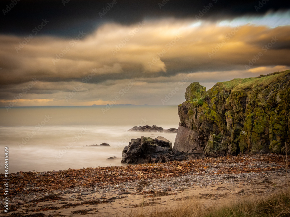 Naklejka premium Machrihanish Beach near Campbeltown.The image is a photograph of a rocky beach with waves under a dramatic, cloudy sky