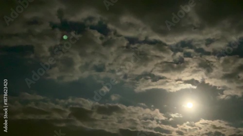A bright full moon in a cloudy night sky. This phenomenon highlights the reflected sunlight from the moon against the dark sky, while the cloud cover softens it.