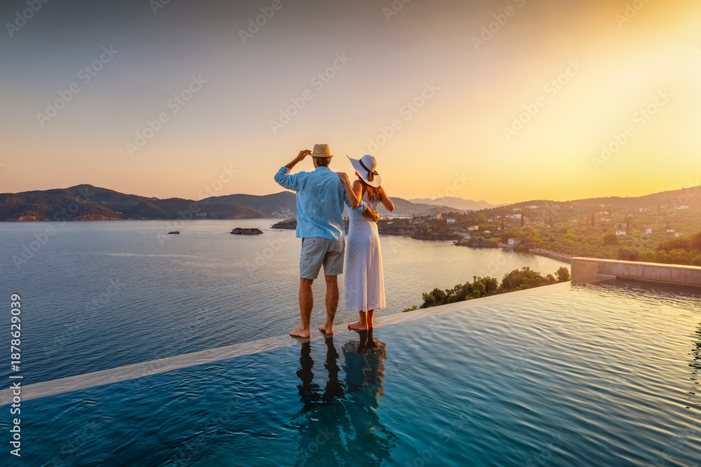 Naklejka premium A romantic couple on summer vacation stands by the infinity swimming pool enjoys a the sunset view behind the ocean