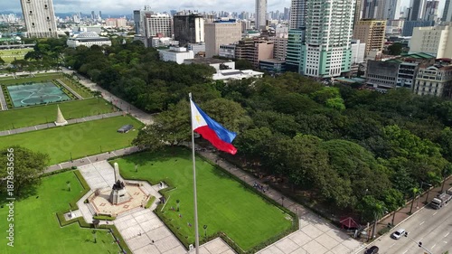 Slow motion aerial view of the Philippine flag waving proudly at Rizal Park in Manila. Urban skyline with high-rise buildings and green spaces frame this patriotic symbol in the heart of the capital.