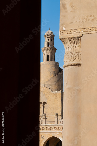 Ibn Tulun Mosque in Cairo viewed through arches. The mosque features a tall minaret and intricate architectural details. Blue sky visible in the background.