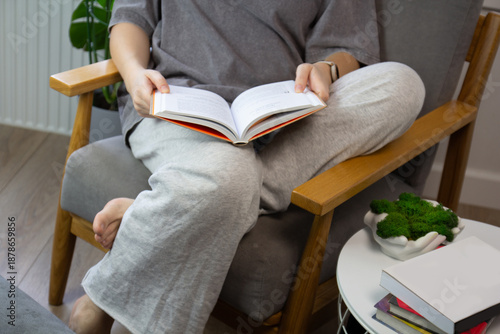 Cropped view of a woman reading a book barefoot in a gray armchair at home. Casual loungewear, soft daylight, and a side table with books create a calm mood