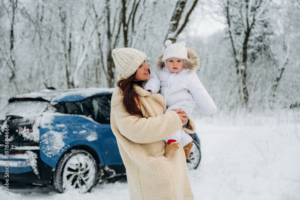 Obraz premium Woman in cozy winter coat holds child in snowy landscape near blue car, showcasing family bonding and joy during winter activities in a serene environment