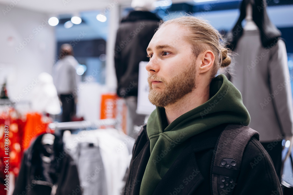 Obraz premium Young bearded man standing in a clothing store, browsing fashion items.