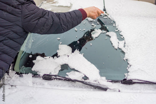 Fotografija a person's hand using a special scraper to clear a layer of melting snow and ice from the hood and windshield of a light-colored car during winter frosts