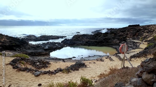 Natural pool formed by lava rock on the sandy coast of Charco del Palo in Lanzarote, Canary Islands, with waves crashing in the background on a cloudy day, creating a unique swimming spot