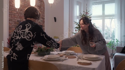 Mother and son setting festive table for Christmas dinner in cozy home interior. Family preparing for holiday celebration near decorated tree with glowing lights and gift boxes.