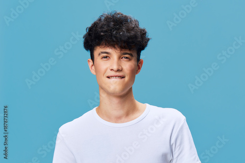Slika na platnu young man with curly hair biting lip posing confidently in white t-shirt against blue background, casual style, playful expression, studio portrait