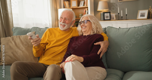 A senior couple sits close together on a couch watching television. They share smiles and seem to enjoy their time together. The living room has warm lighting and a cozy feel.