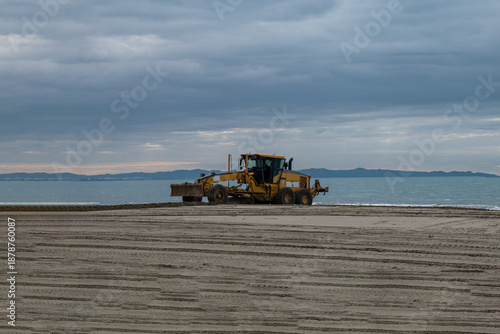 Bulldozer leveling sand on coastal beach near the sea under dramatic cloudy sky, construction machinery, coastal maintenance and environmental management concept.