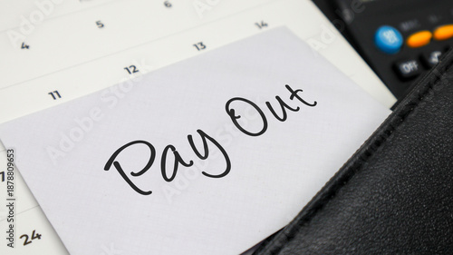 Handwritten words PAY OUT on a white note placed on a calendar with a calculator and leather folder.