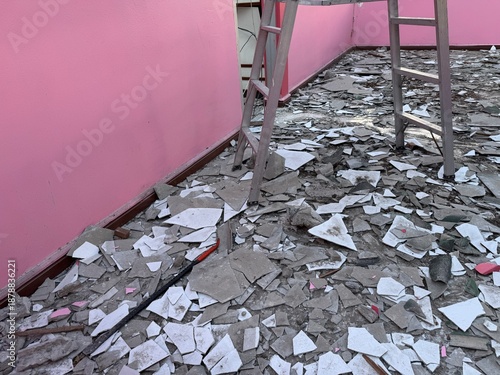 Interior construction site with broken tiles and debris scattered across the floor, a metal ladder standing against a pink wall, showing renovation, demolition mess, and unsafe working conditions.